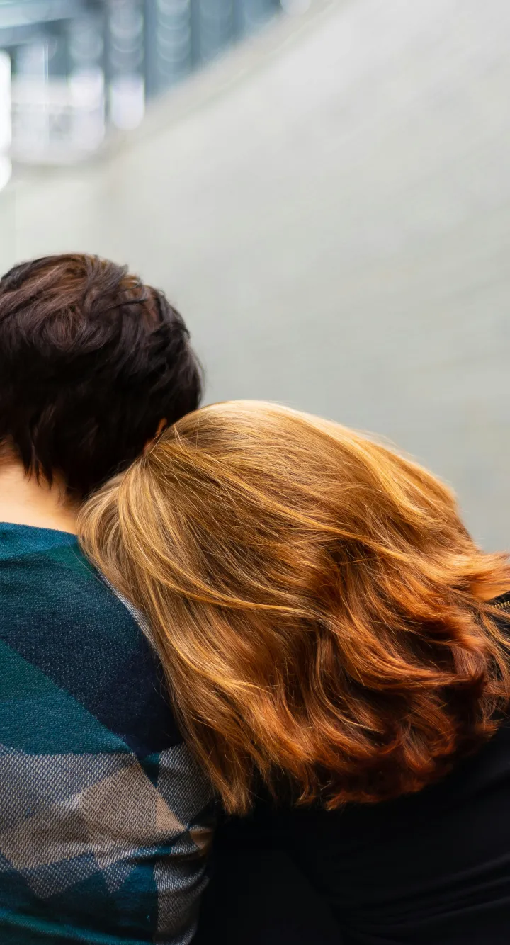 Un couple assis l'un à côté de l'autre dans le hall d'un hôpital. La femme aux cheveux roux pose sa tête sur l'épaule de son compagnon.