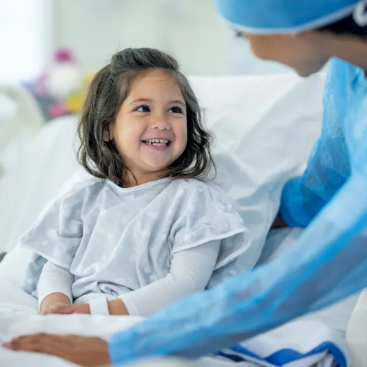 Petite fille dans son lit d'hôpital, souriant à l'infirmière qui s'occupe d'elle.
