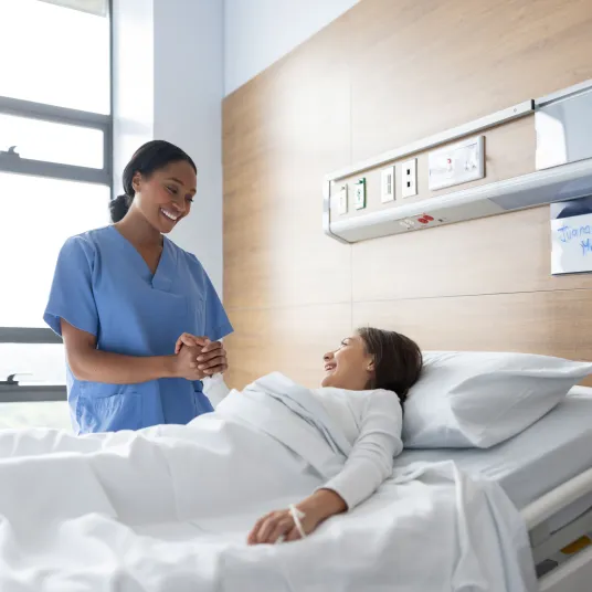 Une infirmière tenant la main d'une patiente alitée et la regarde avec un sourire. groupe Vivalto Santé