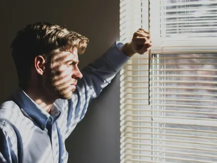 Un homme l'air perturbé, regarde par la fenêtre de sa maison.